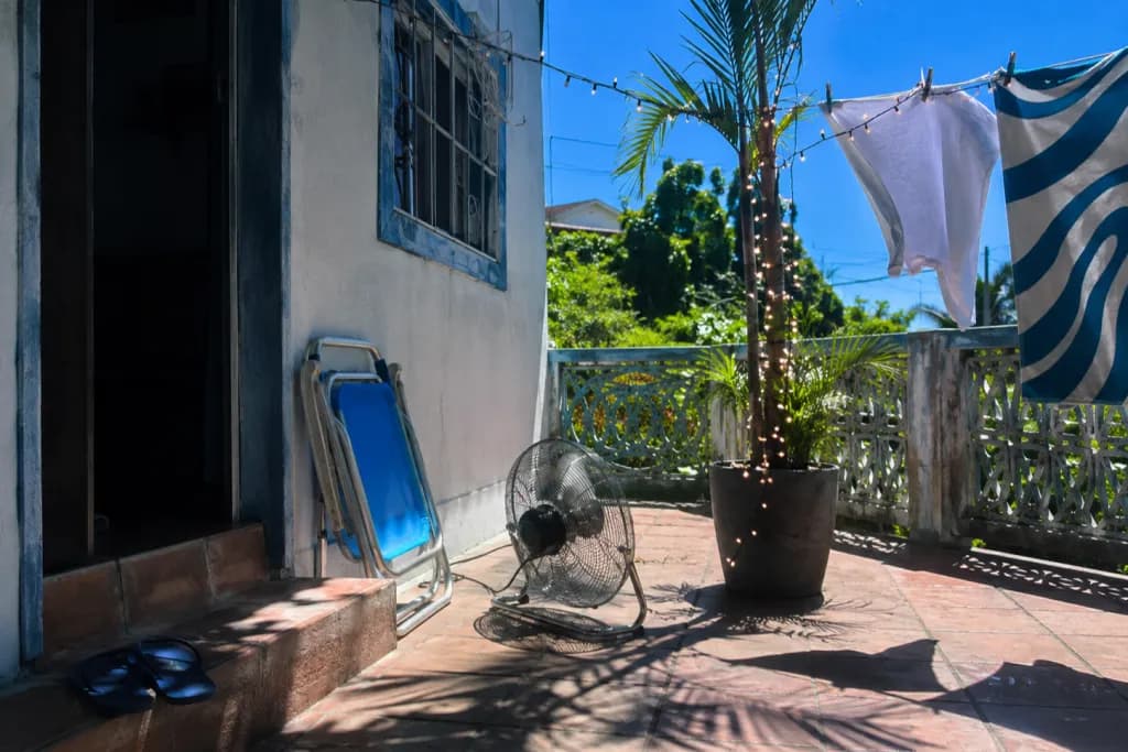 Varanda brasileira ao sol de dezembro: palmeira em vaso com luzes de Natal e bolas, varal com roupa branca, ventilador de chão e cadeira de praia dobrada