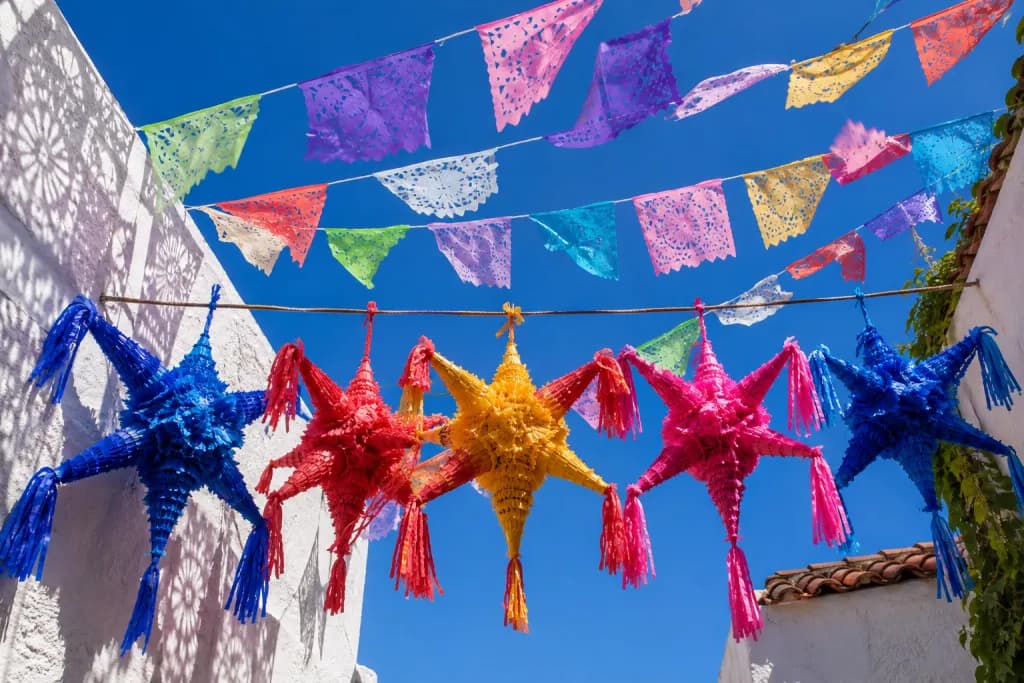 Piñatas de siete picos y papel picado de colores colgados sobre un patio mexicano encalado, contra un cielo azul de mediodía